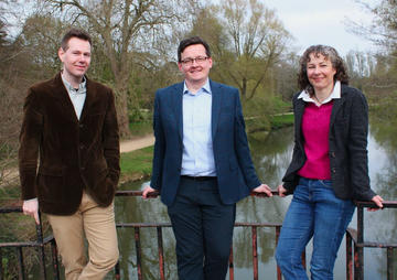Three people wearing jackets and shirts stand on a bridge in Oxford's University Parks.