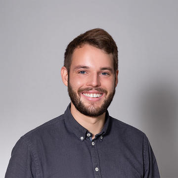 Portrait photo of Andy Sode Anker, a smiling man with brown hair and a beard who is wearing a navy button-up shirt standing against a grey background.