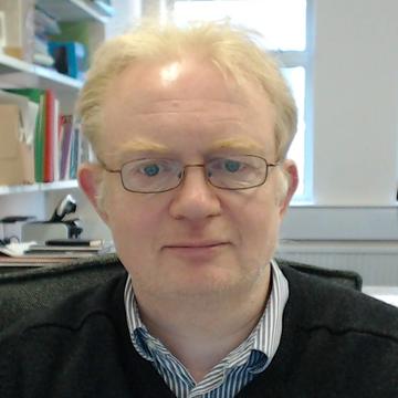Headshot photograph of Martin Owen Jones, a man with light hair and glasses wearing a shirt and black jumper, sat in an office.