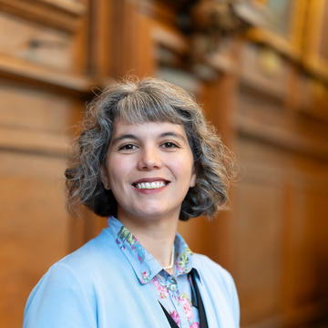 A woman smiling with curly grey hair wearing a blue cardigan and a pink and blue blouse.